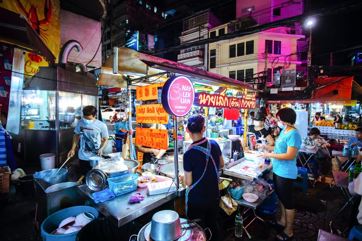 Bustling night market food stall with vibrant signage, people cooking and serving dishes under neon lights.