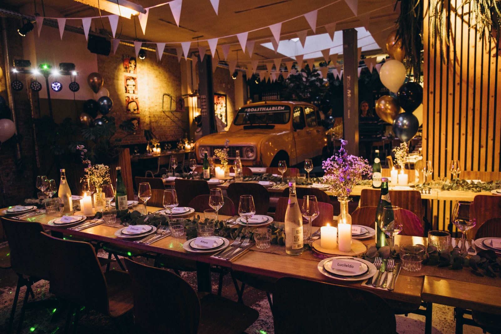 Gezellige feestelijke tafelopstelling met kaarsen, bloemen en servies, omgeven door sfeervolle verlichting en een gele auto.