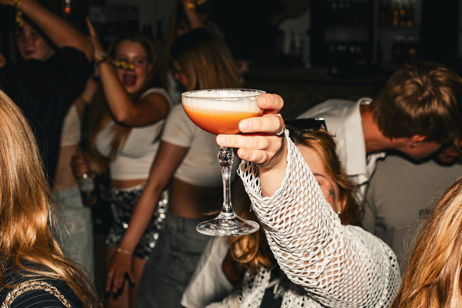 Hand houdt een kleurrijke cocktail omhoog in een drukke bar met feestende mensen op de achtergrond.
