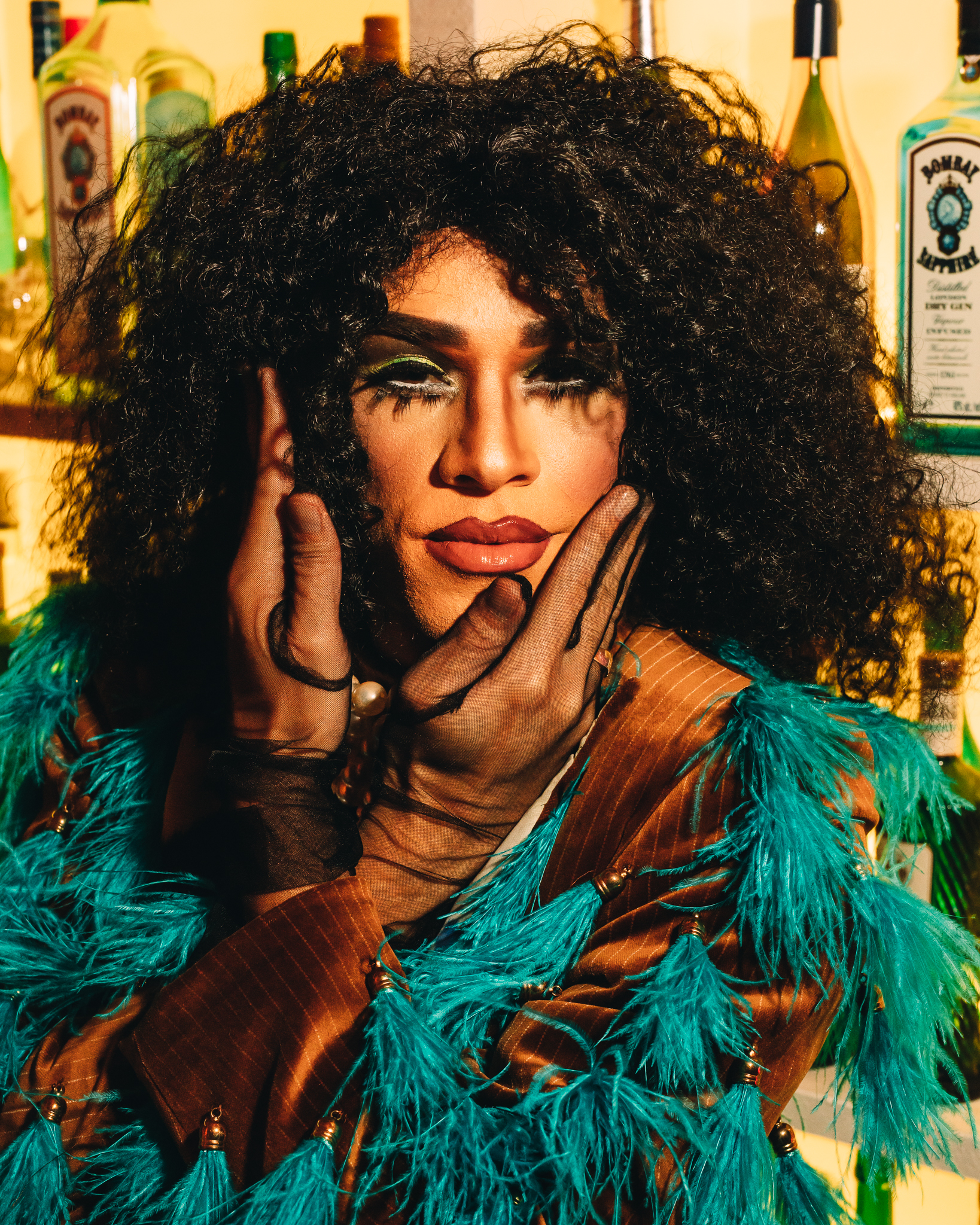 A dragqueen with dramatic makeup and curly hair poses with hands on their face, wearing a teal feathered outfit, in front of a shelf of liquor bottles.