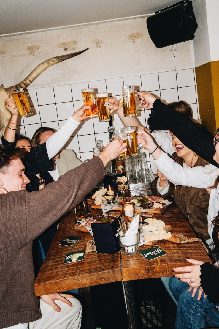 Gezellig gezelschap proost met bierglazen rond een houten tafel vol hapjes in een sfeervol café.