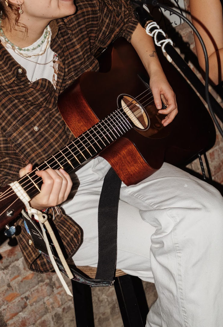 Persoon speelt akoestische gitaar, gekleed in geruite blouse en witte broek, tegen bakstenen muur.
