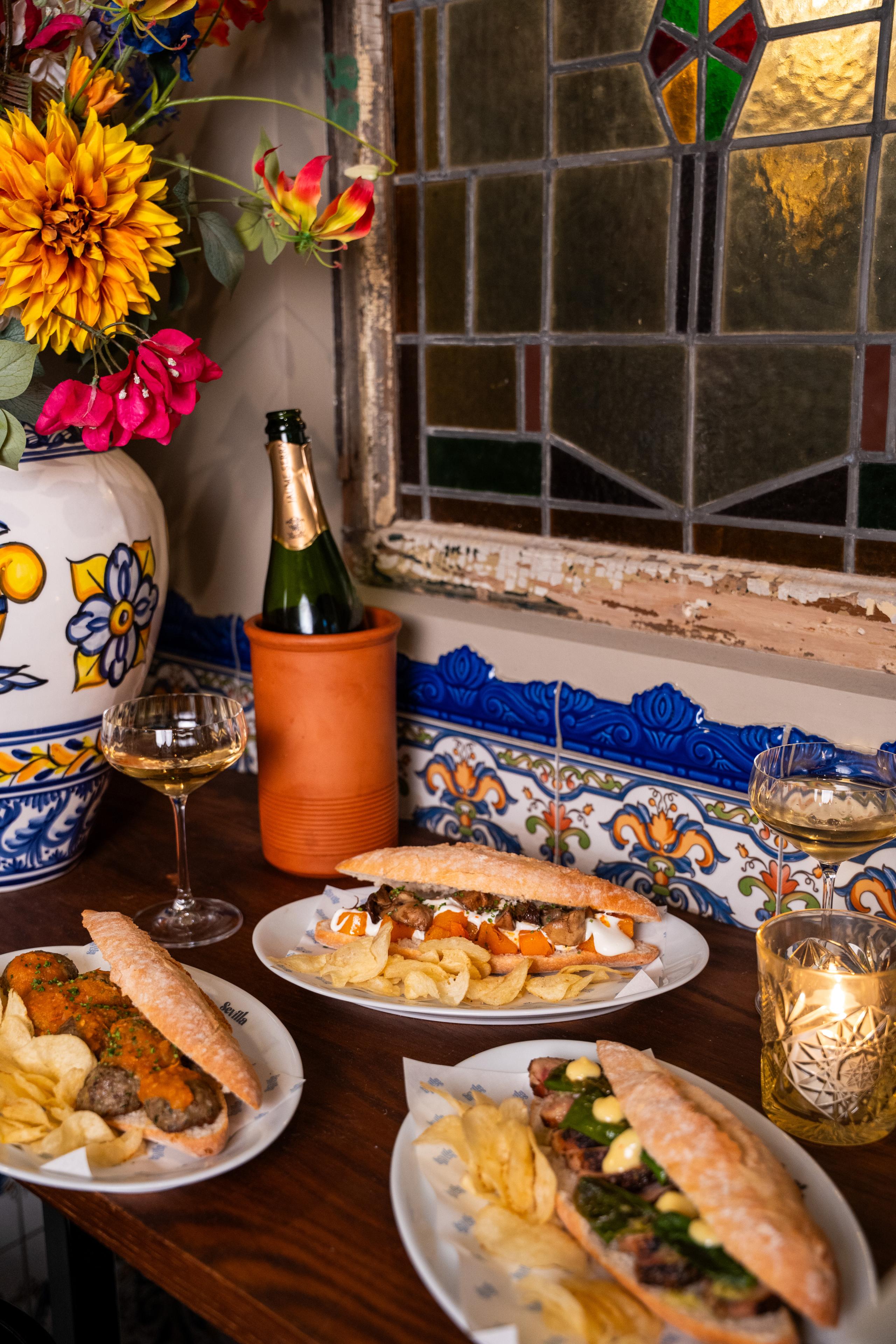 Drie baguettes met chips op houten tafel, kleurrijk bloemenvaas, glas champagne, sfeervol glas-in-loodraam.