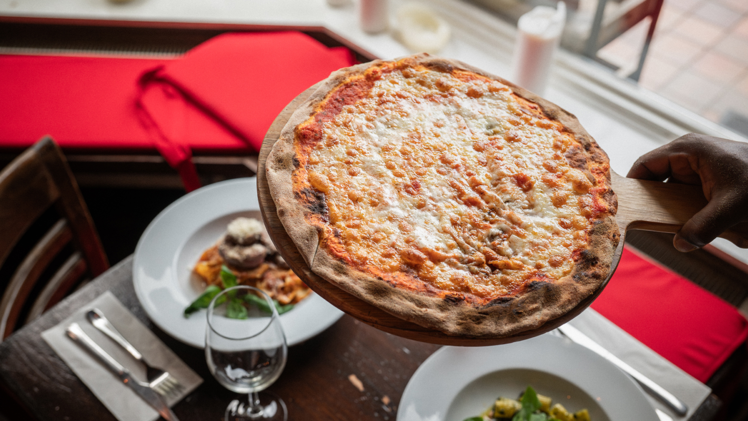 Freshly baked margherita pizza with golden crust, held above a table set with pasta dishes and red seating.