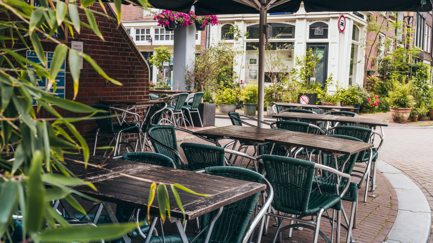 Outdoor cafe with green wicker chairs and wooden tables, surrounded by lush plants and brick walls.