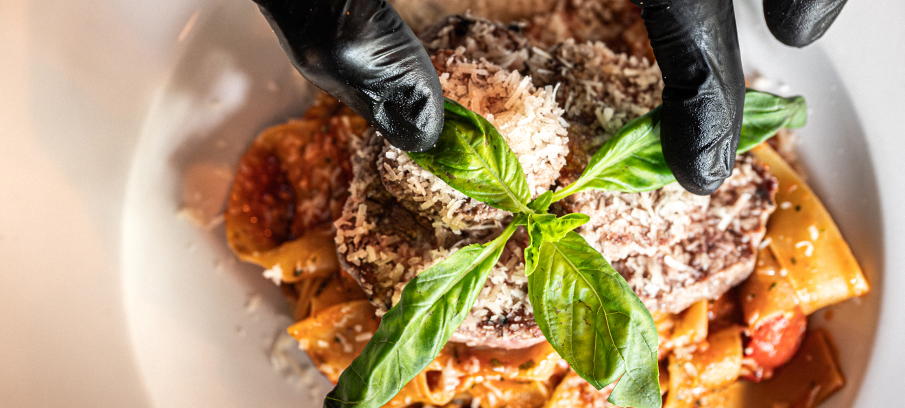Gloved hands garnishing pasta with fresh basil atop a rich tomato sauce, sprinkled with Parmesan cheese.