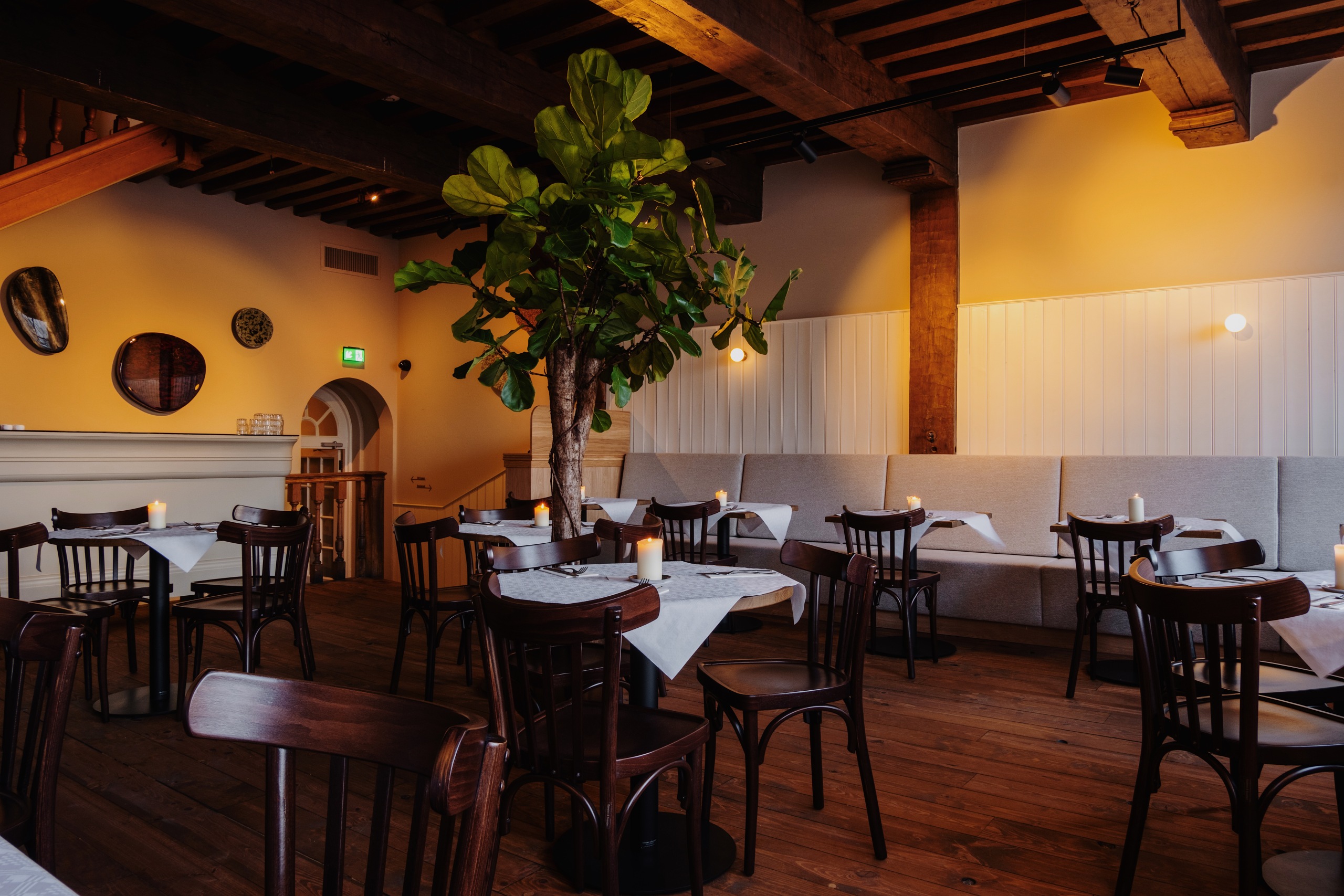 Gezellige restaurantinterieur met houten tafels, kaarslicht, groene plant en warme verlichting.