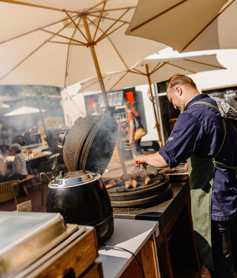 Chef grilt worstjes op een barbecue onder grote parasols, ontspannen terras met gasten op de achtergrond.