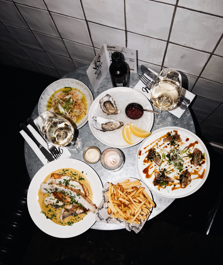 Ronde tafel met oesters, visgerecht, friet en witte wijn, sfeervol verlicht in een moderne eetomgeving.
