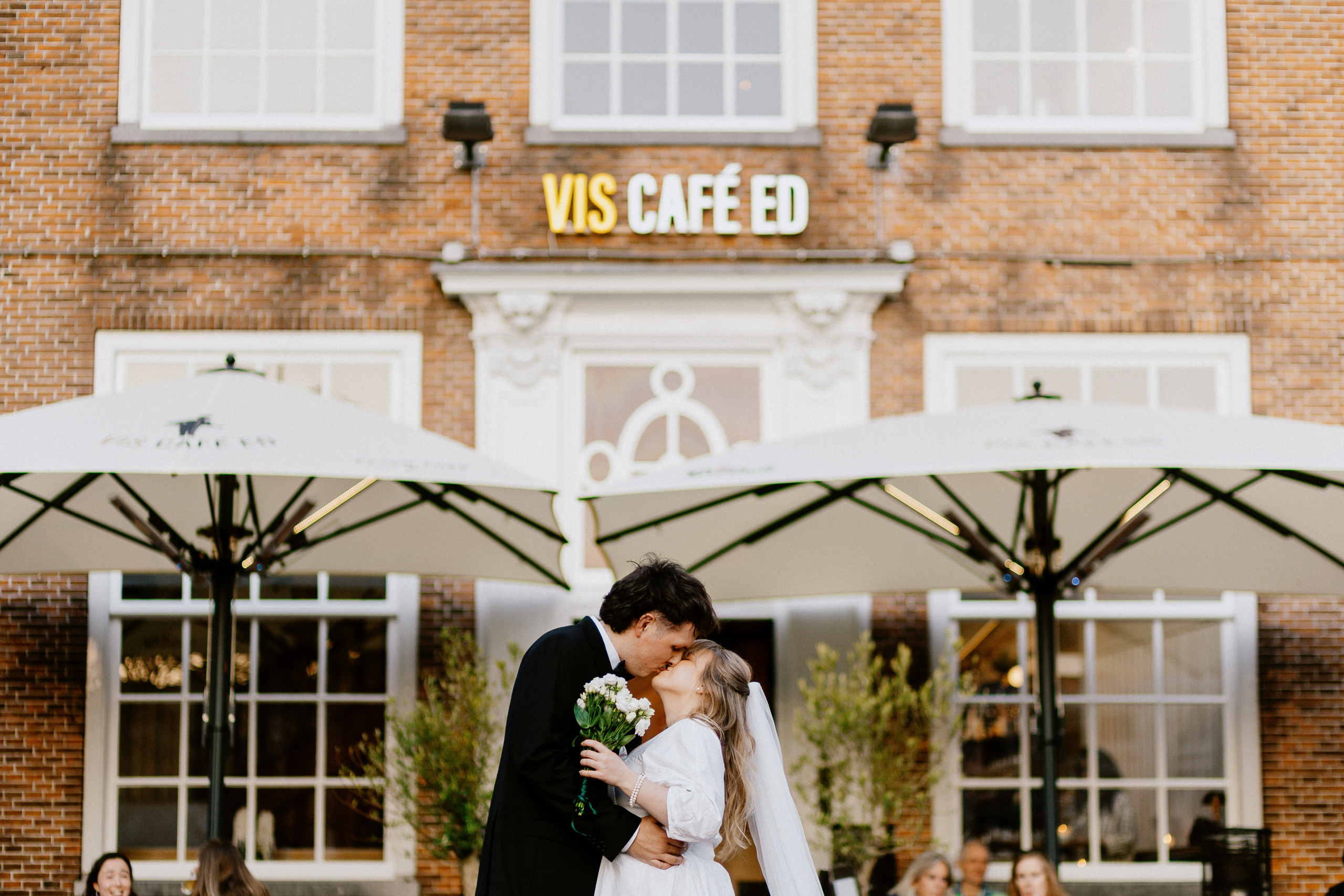 Bruidspaar kust onder parasols voor Vis Café ED, met bakstenen gevel op achtergrond.