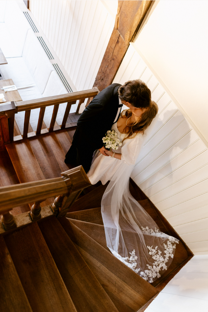 Bruidspaar op houten trap, bruid in witte jurk met bloemen, warme houten en witte wanddecoratie.
