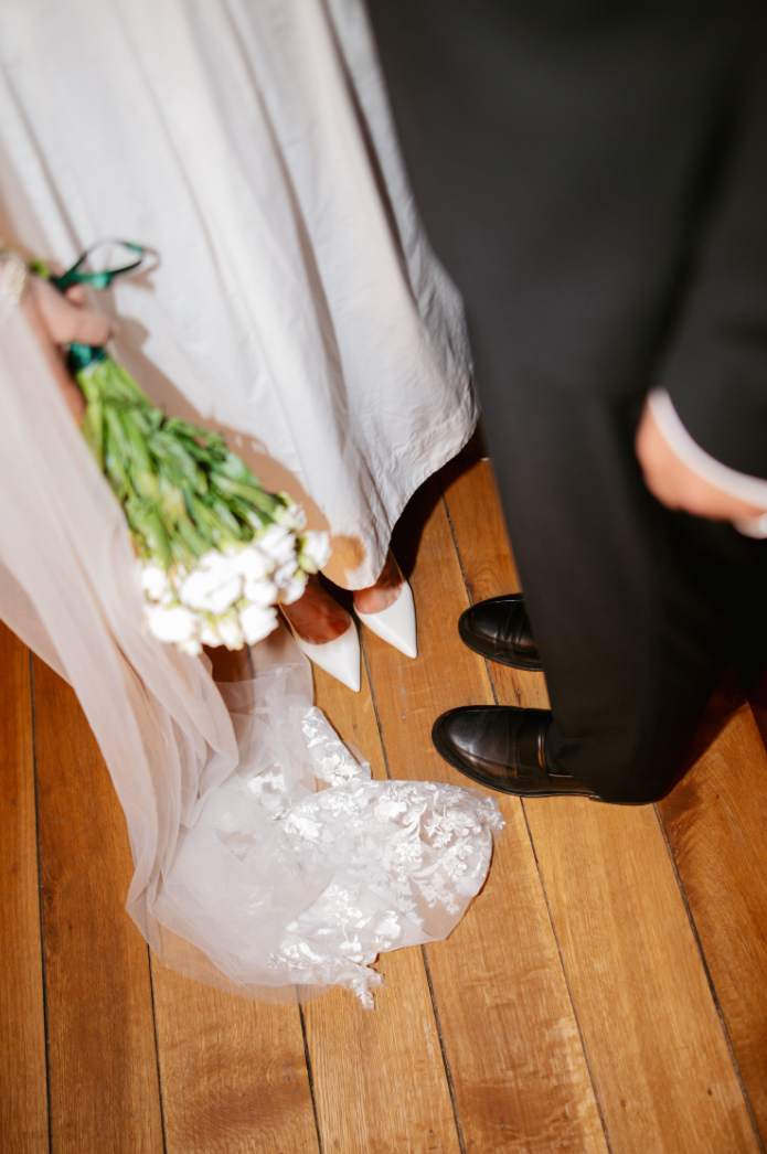 Bruidsjurk met kanten sleep en witte schoenen naast zwarte herenschoenen op houten vloer.