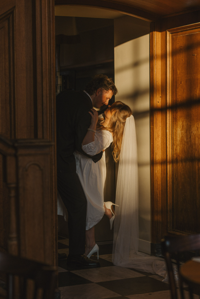 Bruidspaar in romantische omarming bij zonsondergang, warm licht valt op houten wand en zwart-wit geblokte vloer.