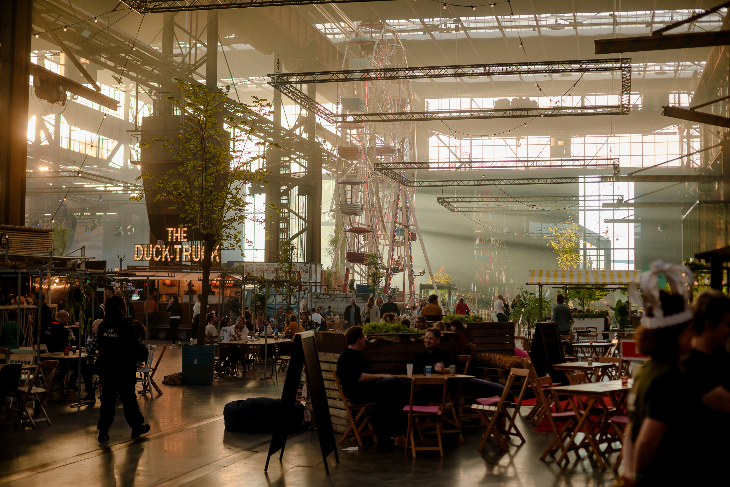 Industriële hal met foodtrucks, terrasstoelen en een reuzenrad. Zachte verlichting en levendige sfeer.