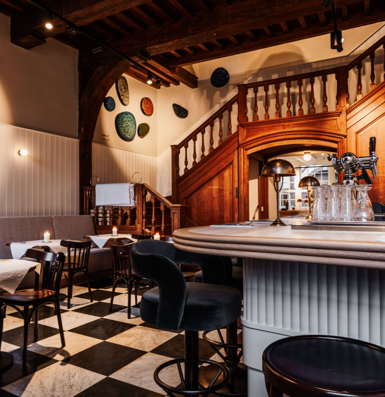 Gezellige restaurantinterieur met houten trap, zwart-wit geblokte vloer, kaarsverlichte tafels en bar.