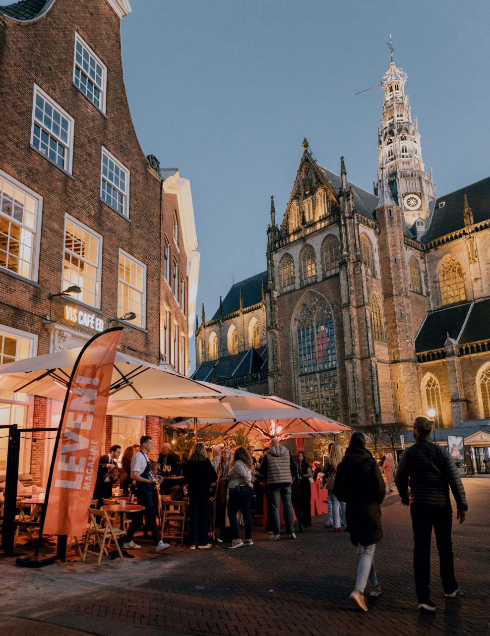 Terras van café in avondlicht, mensen genieten onder witte parasols, historische kerk op achtergrond.