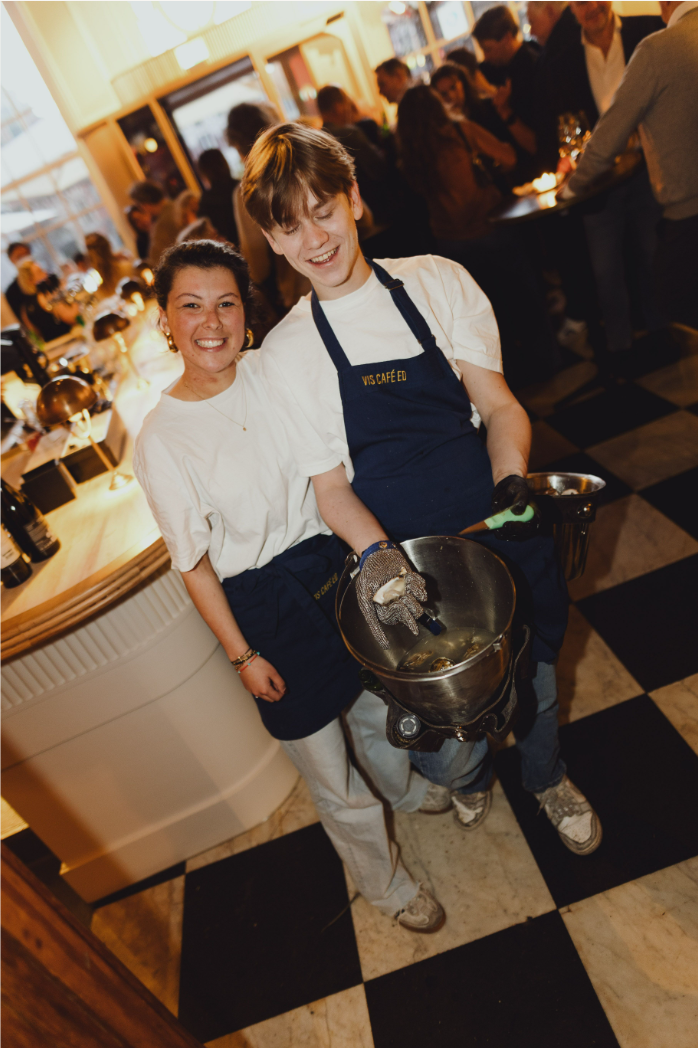 Twee lachende medewerkers in een drukke restaurantomgeving, met marmeren vloer en warme verlichting.