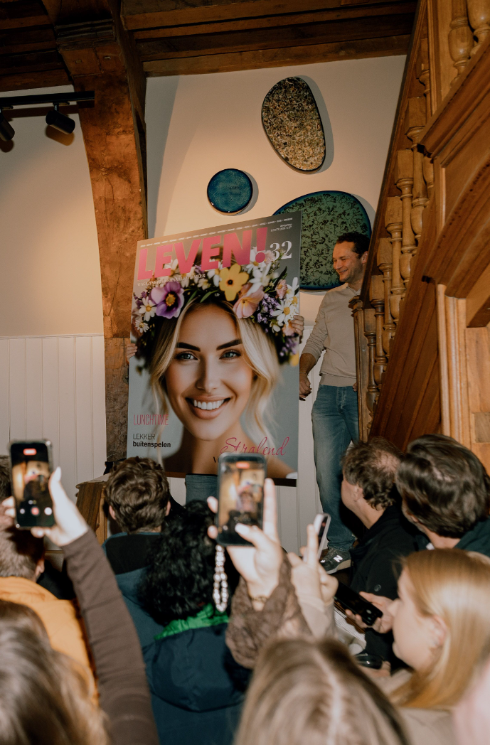 Tijdschriftcover met bloemenkrans, omringd door mensen die foto's maken in een houten interieur.