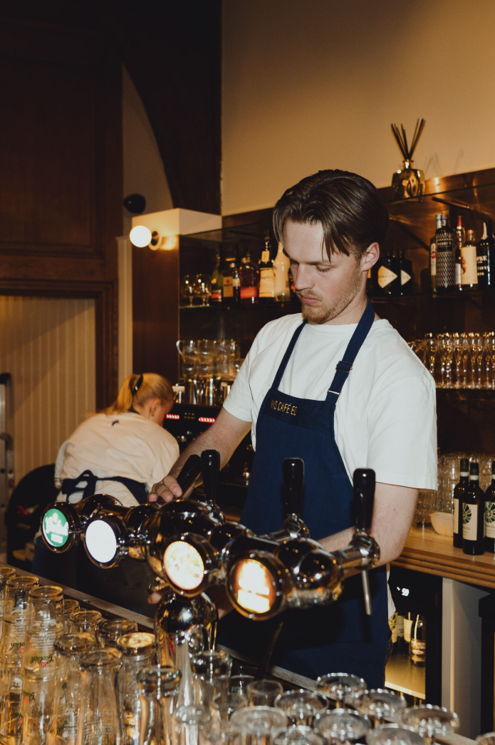 Barman in blauw schort tapt bier in een sfeervolle bar met houten accenten en glazen op de toonbank.