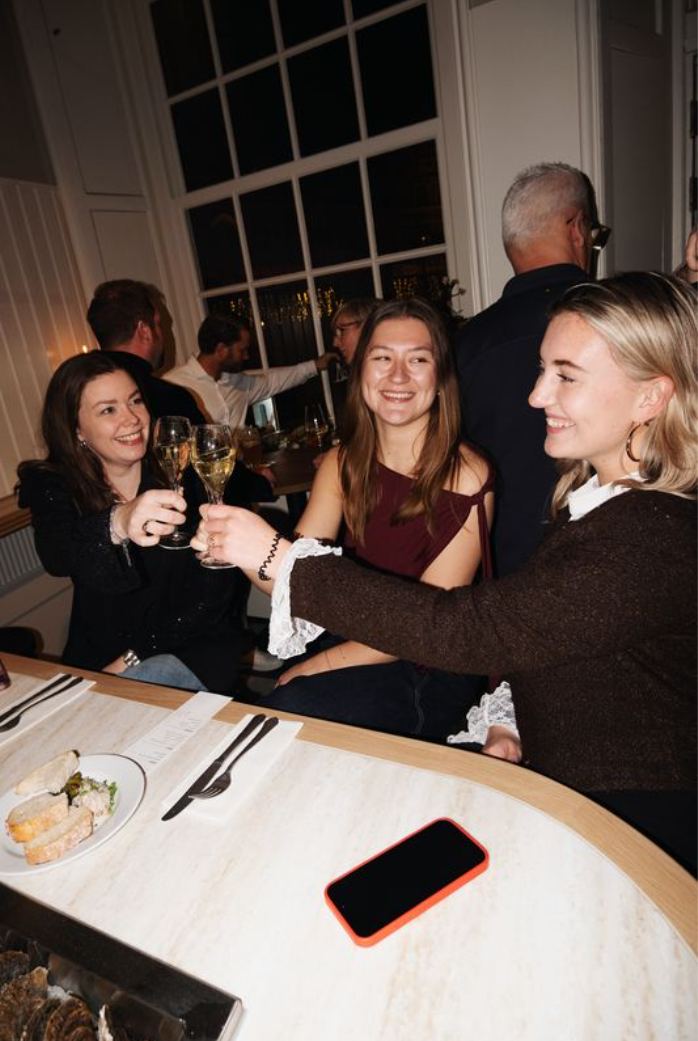 Drie vrouwen proosten met champagne in een gezellige restaurantsetting, omringd door andere gasten.
