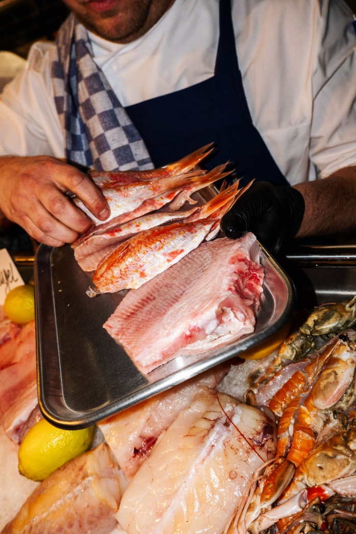 Verse vis op metalen schaal, chef in wit uniform en blauwe schort, omgeven door zeevruchten en citroenen.