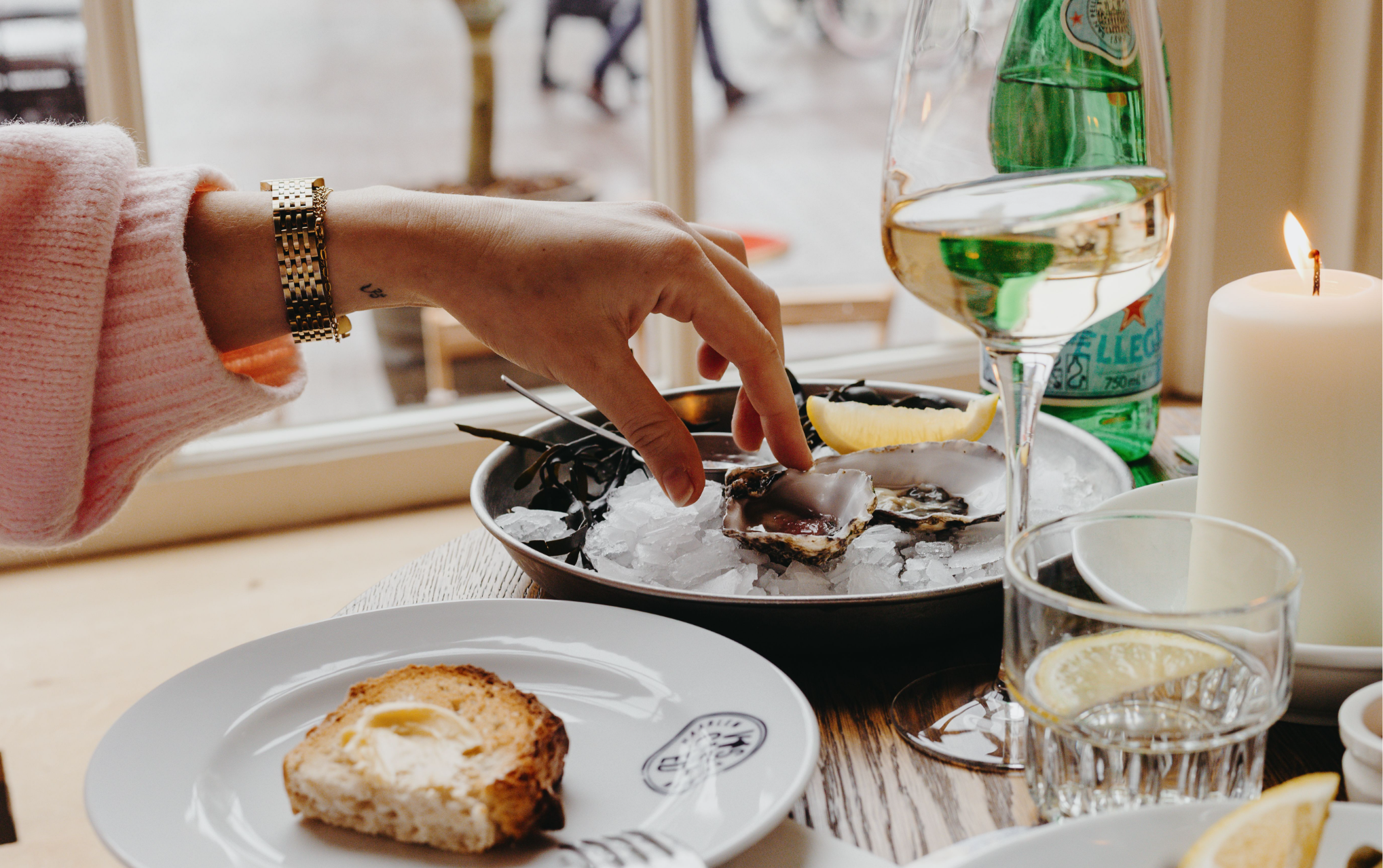 Hand pakt oester van schaal met ijs, vergezeld door glas witte wijn, kaars en broodje met boter.