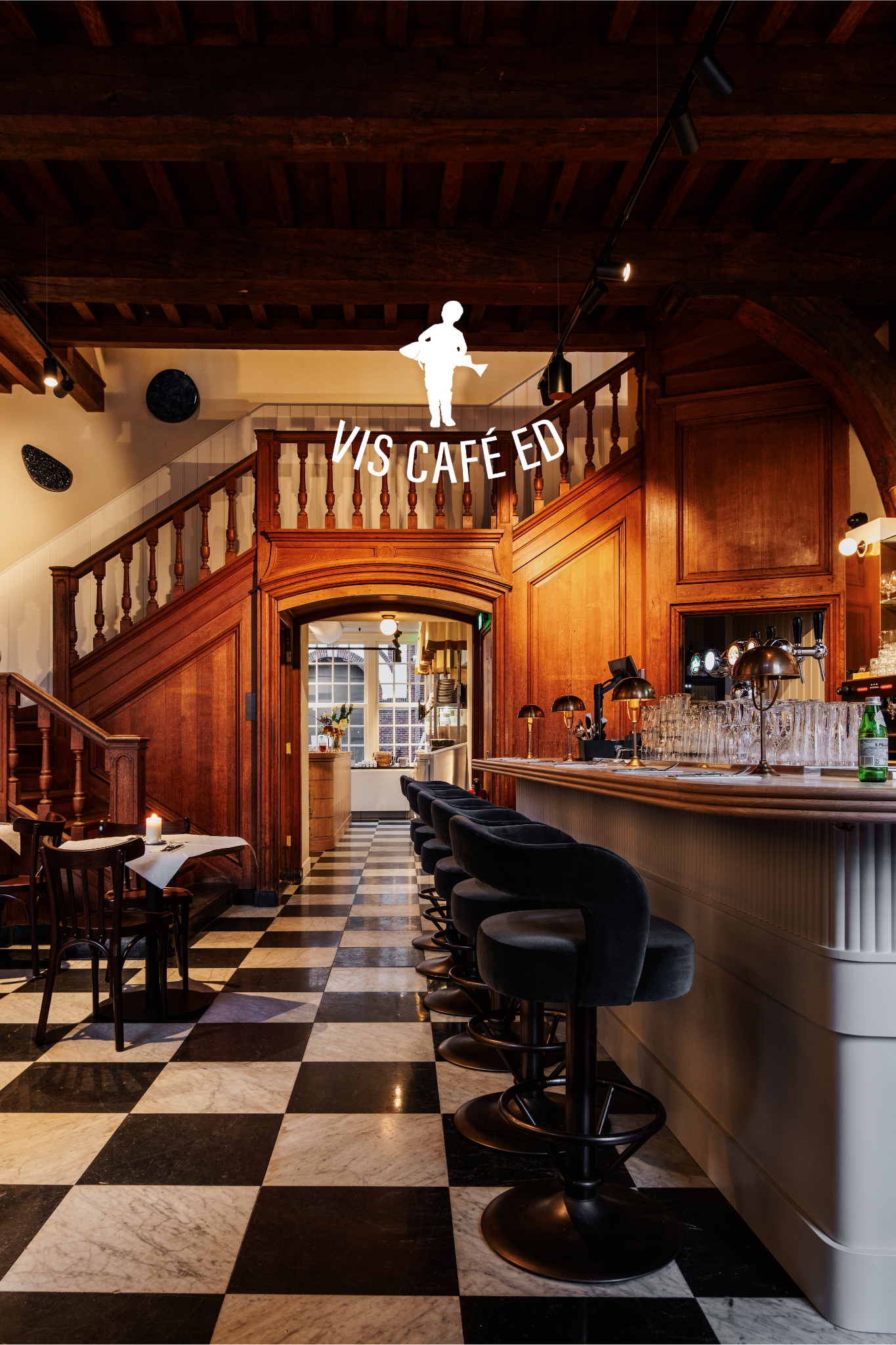 Gezellig café-interieur met houten trap, zwart-wit geblokte vloer, bar met fluwelen krukken en warme verlichting.