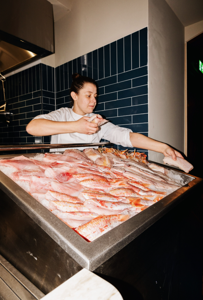 Kok in witte jas schikt verse vis op ijs, tegen een achtergrond van blauwe tegels.