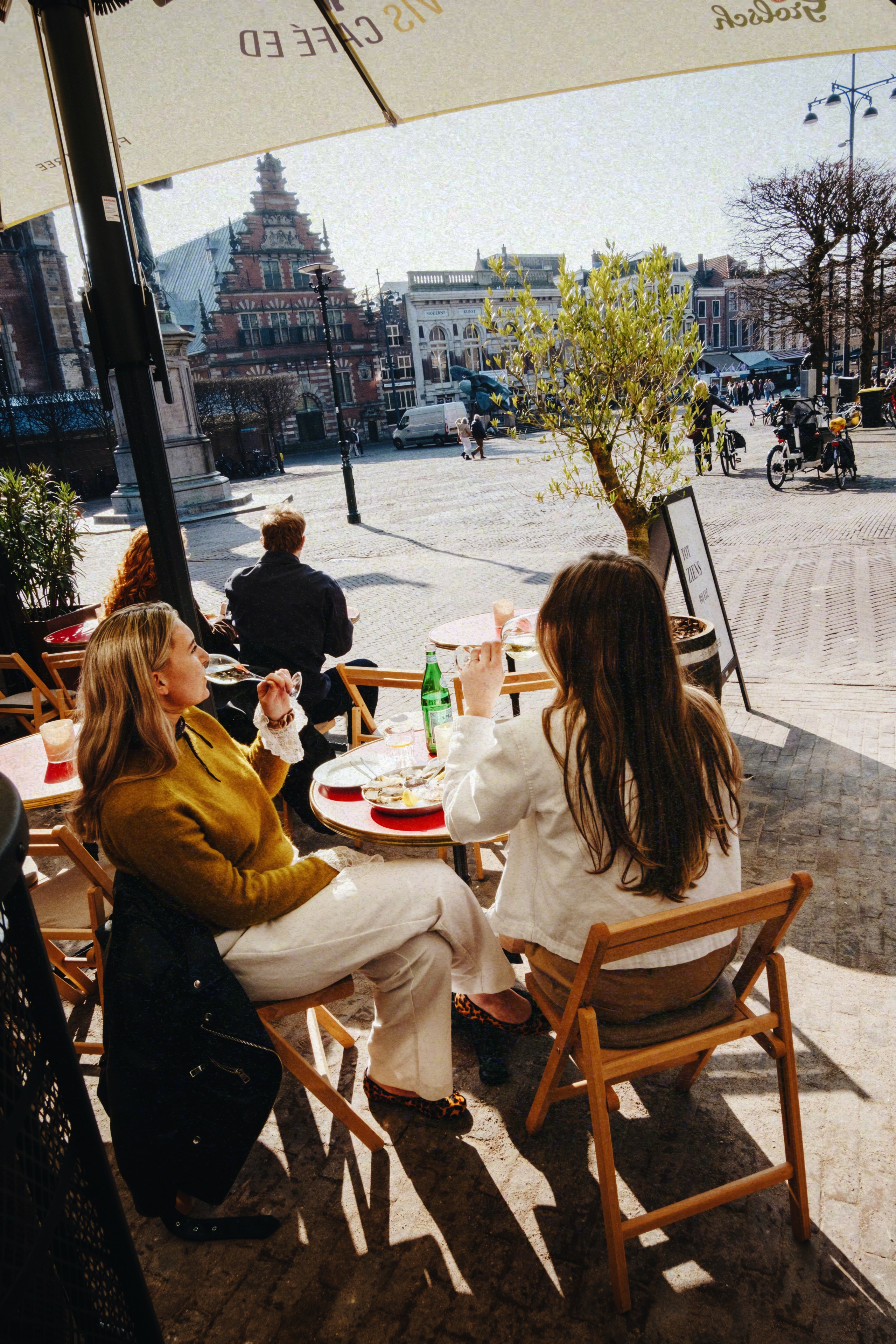 Twee vrouwen genieten van drankjes op een zonnig terras met uitzicht op een historisch plein.