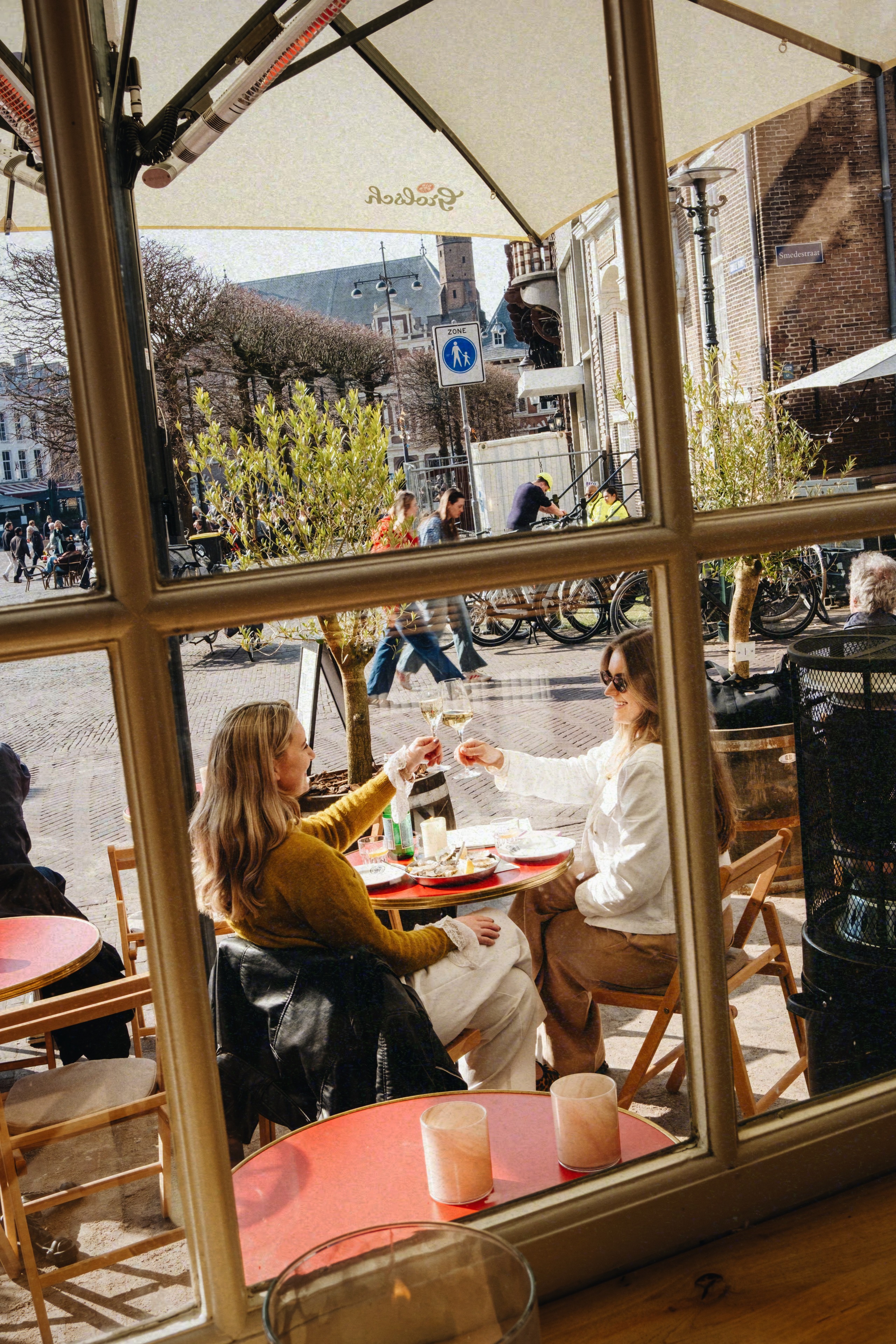 Twee vrouwen proosten met wijn op een zonnig terras met rode tafels, omringd door stedelijke drukte.