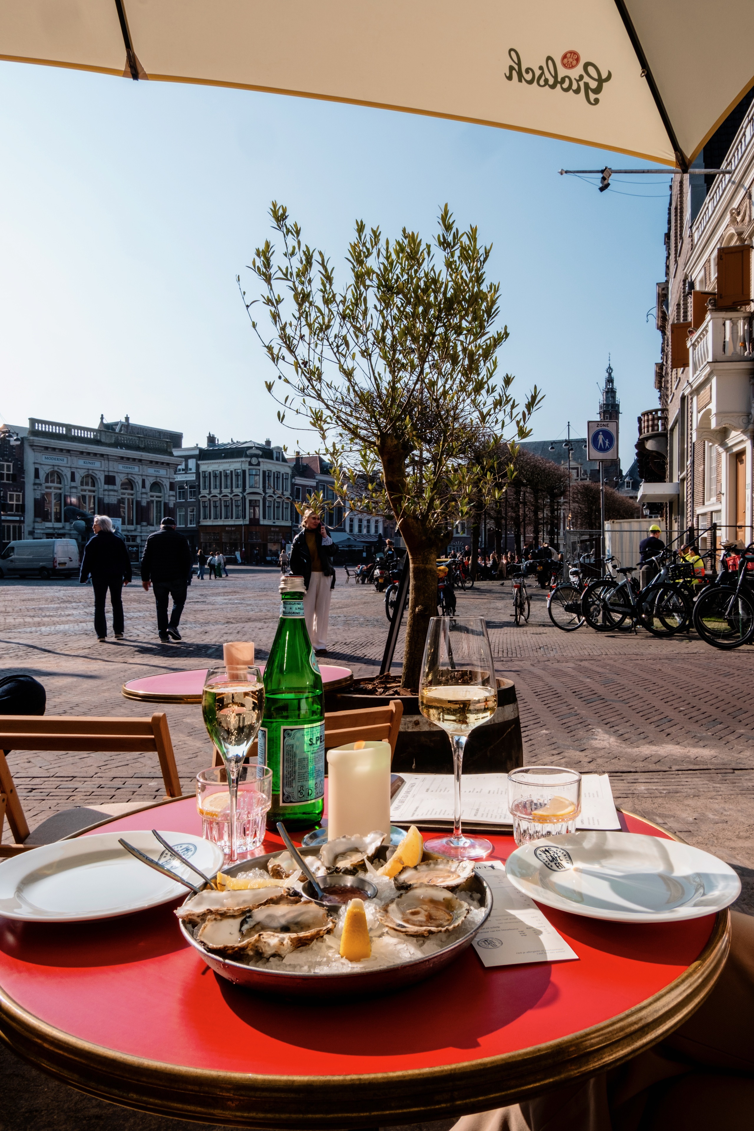 Oesters op ijs met citroen op een rood tafeltje, omringd door glazen wijn, tegen een stedelijke achtergrond.