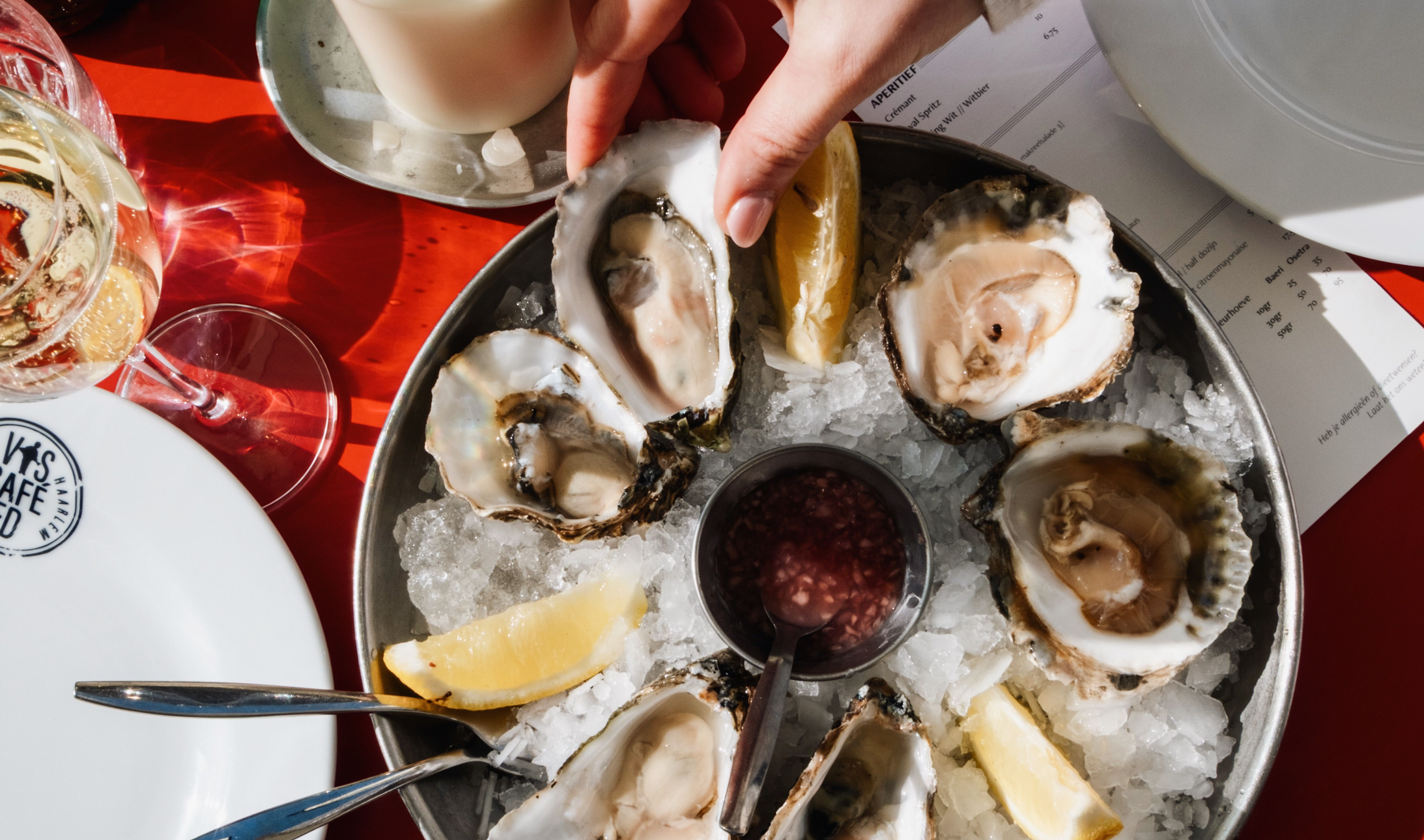 Verse oesters op ijs met citroenpartjes, een hand die een oester pakt, rode tafel, glas witte wijn.