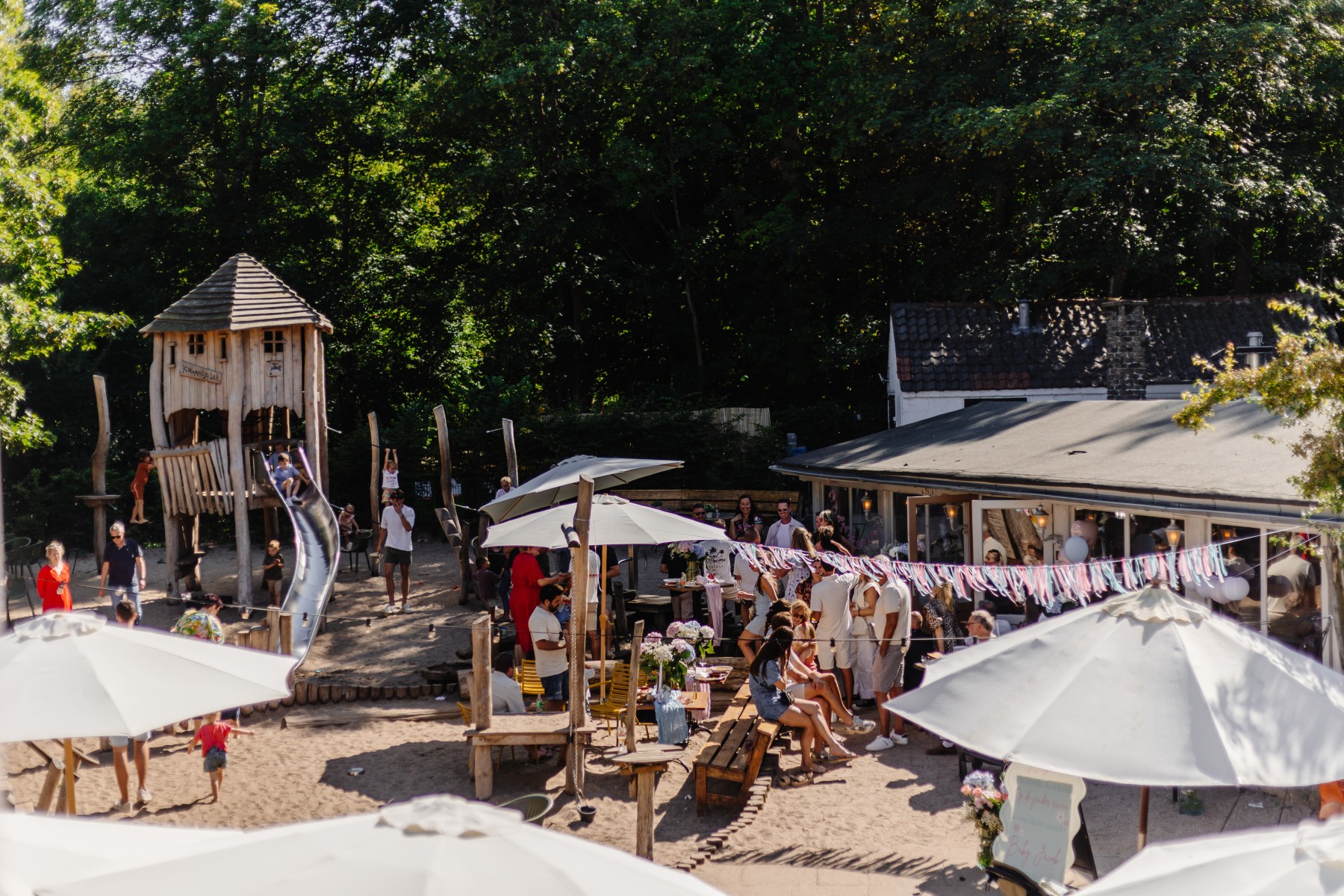 Gezellige speeltuin met houten klimtoestel en glijbaan, omringd door terras met parasols en feestelijke slingers.