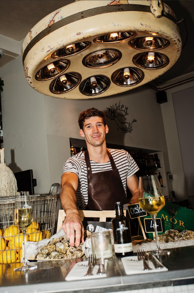 Man in gestreept shirt en leren schort presenteert oesters op een bar. Sfeervolle verlichting en wijn.
