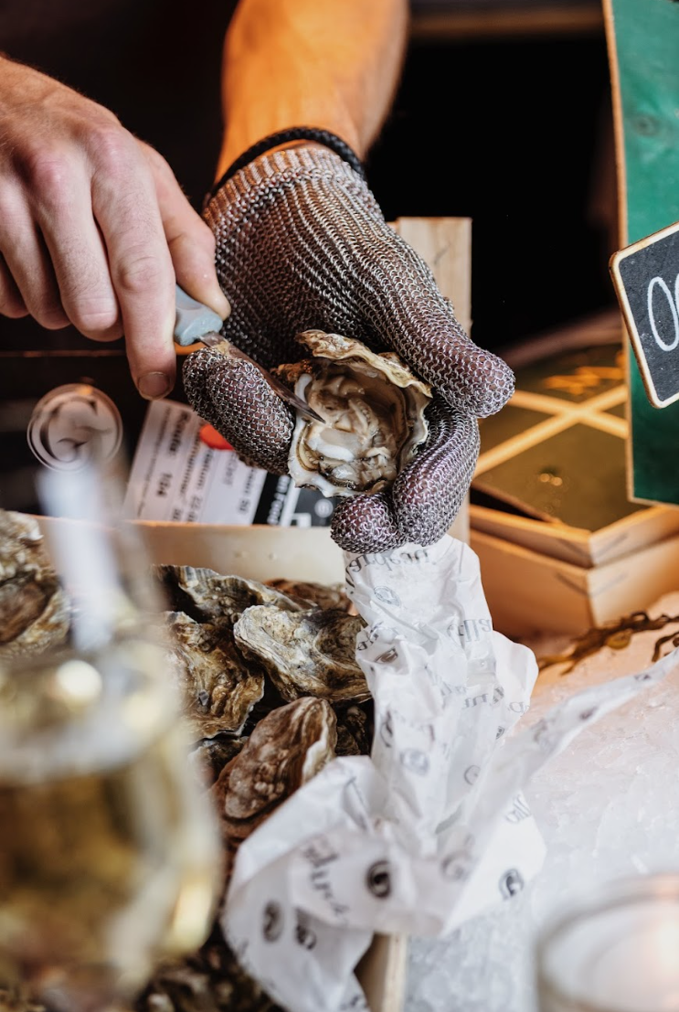 Hand opent oester met handschoen, verse oesters op ijs, sfeervolle marktomgeving op achtergrond.