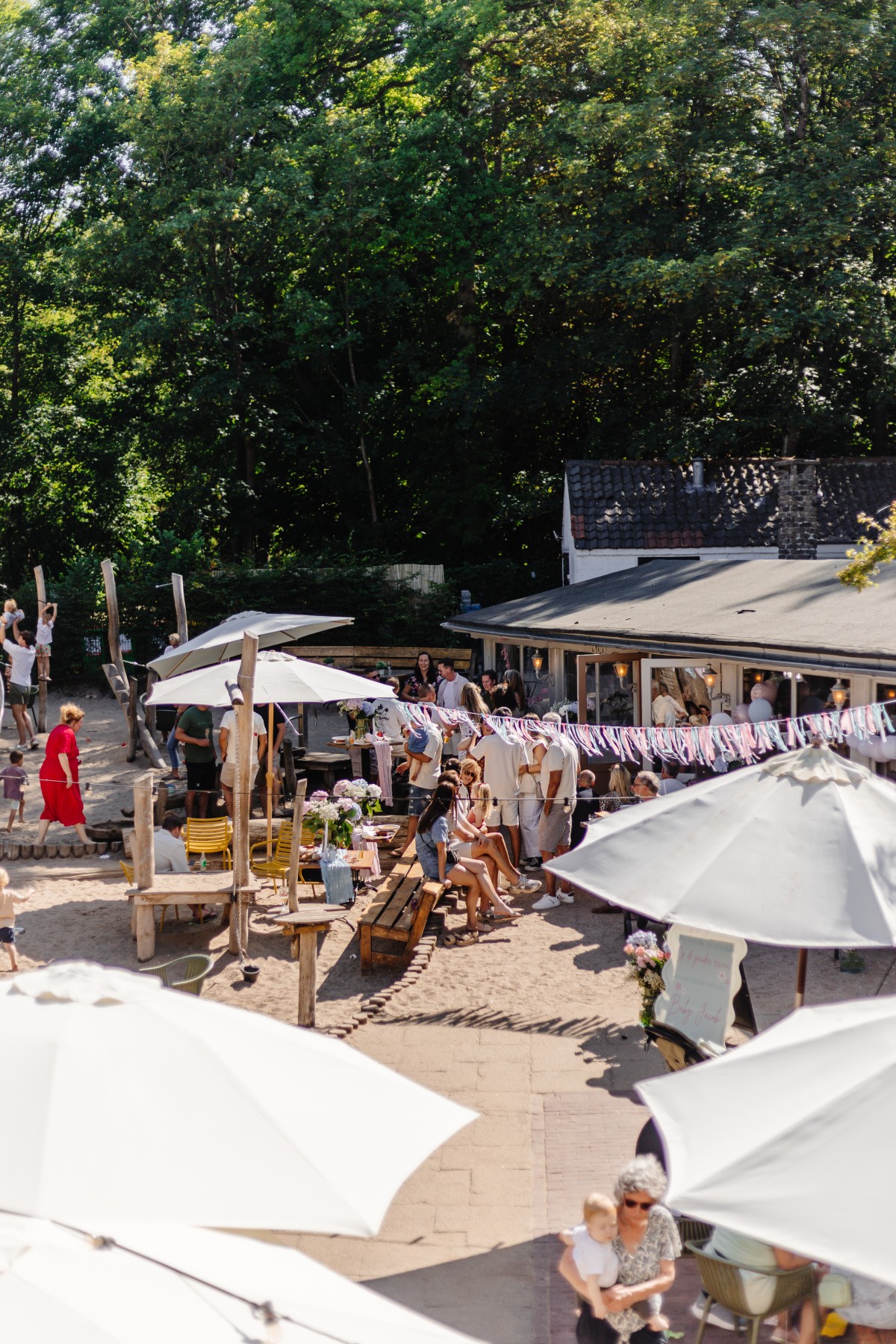 Zonnig terras met houten banken, witte parasols en feestelijke slingers, omringd door groene bomen.