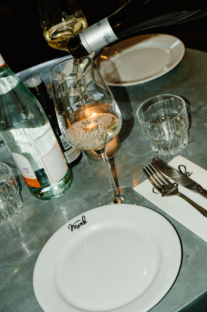 Witte wijn wordt ingeschonken in een glas op een gedekte tafel met een bord, bestek en waterfles.