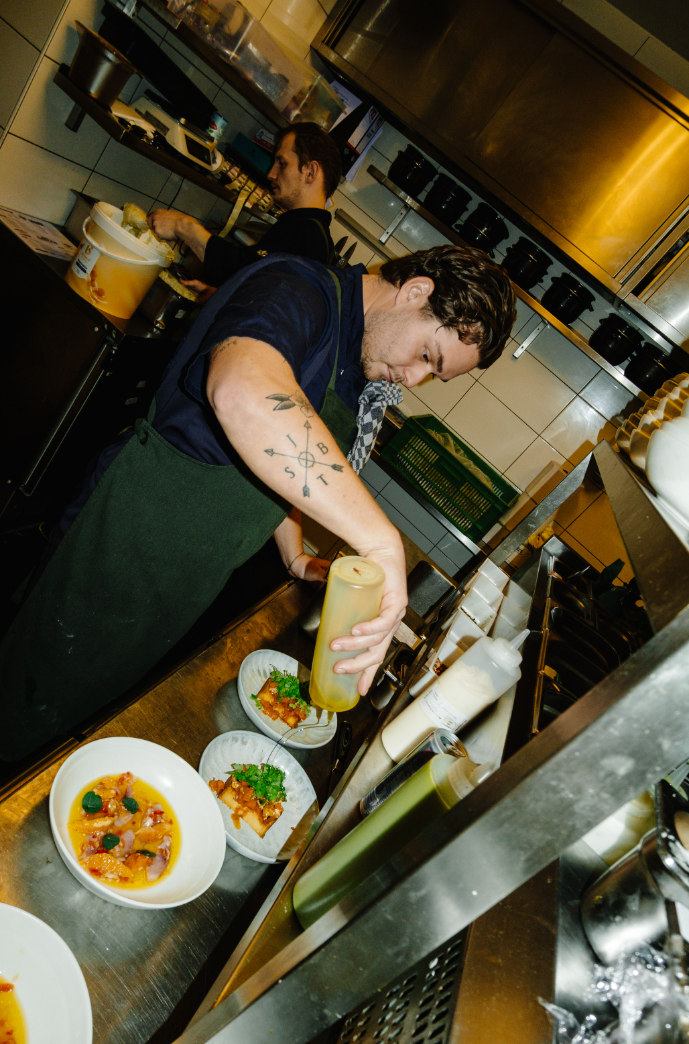 Kok in drukke keuken giet saus over kleurrijke gerechten in witte kommen, omringd door keukengerei.
