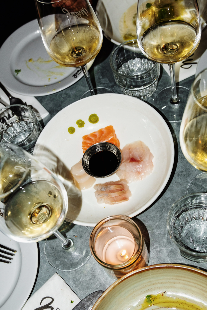 Sashimi op wit bord met sojasaus, omringd door wijnglazen en kaarslicht op een metalen tafel.