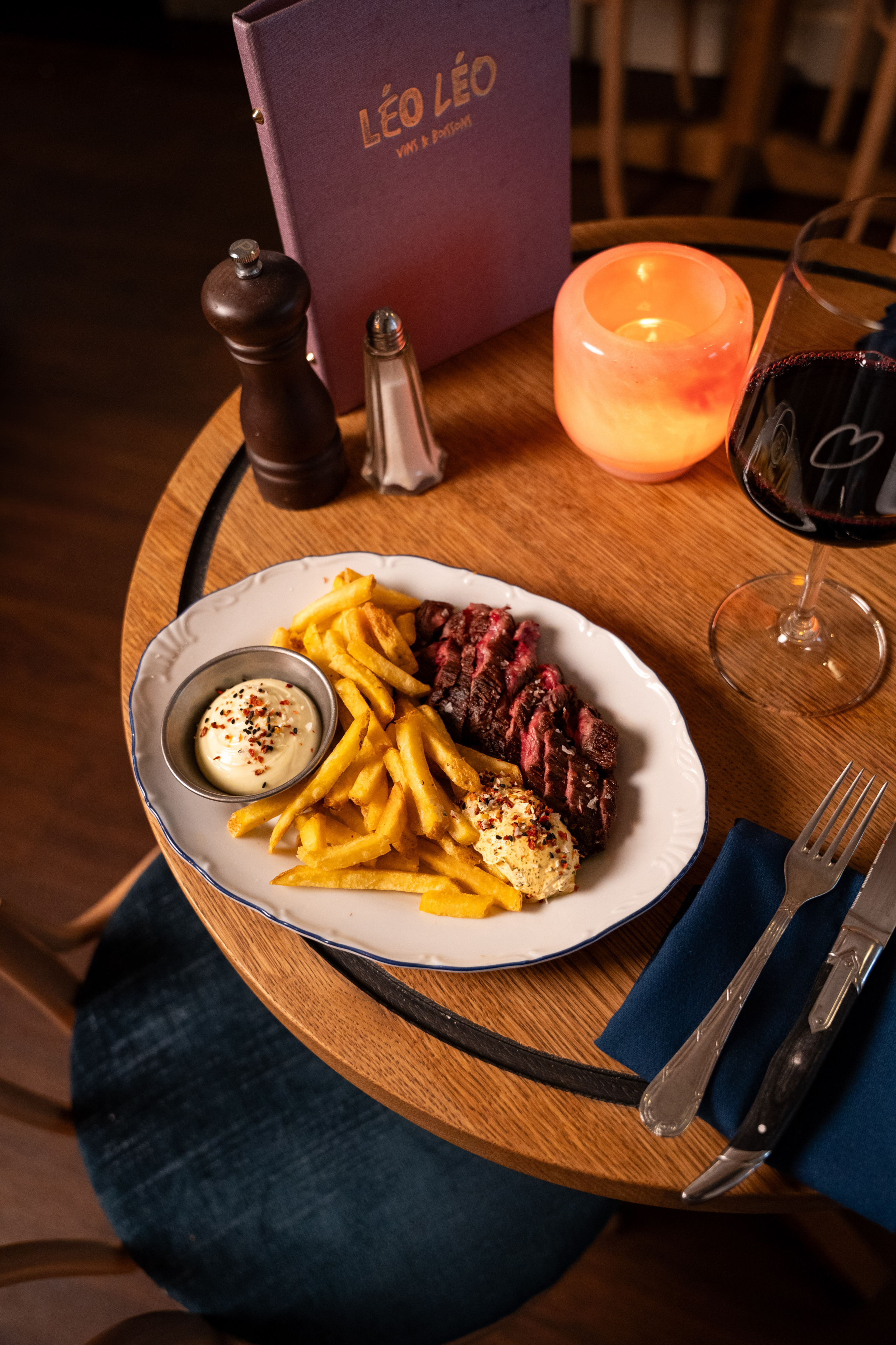 Entrecôte met frietjes en saus naast kaarslicht en een glas rode wijn op een houten tafel.