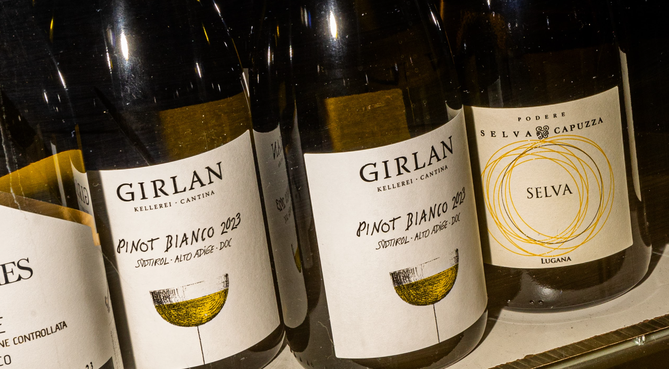 Wijnflessen met etiketten van Girlan Pinot Bianco 2023 en Selva Capuzza, sfeervol verlicht.
