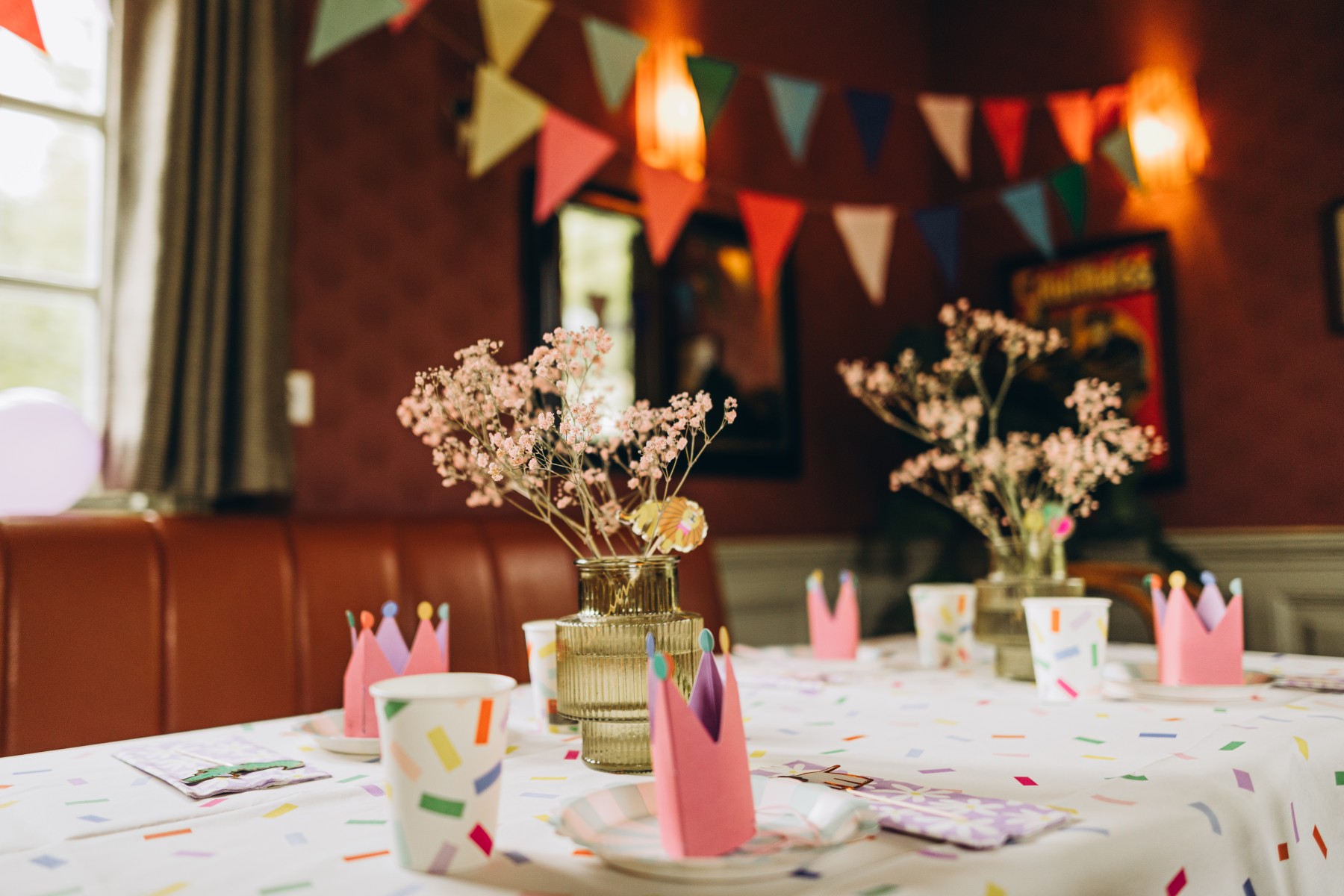 Feestelijke tafel met kleurrijke slingers, roze papieren hoedjes en bloemen in een gezellige ruimte.