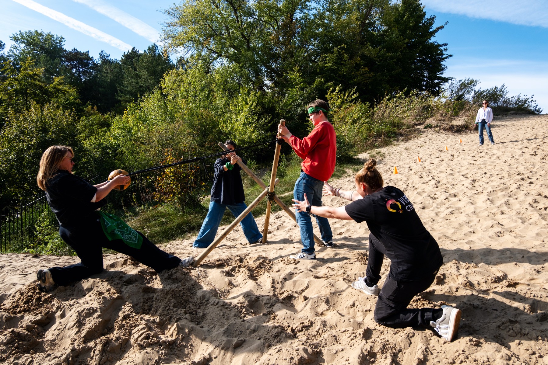 Groep mensen op zandheuvel bezig met teamactiviteit, omringd door groen bos en blauwe lucht.