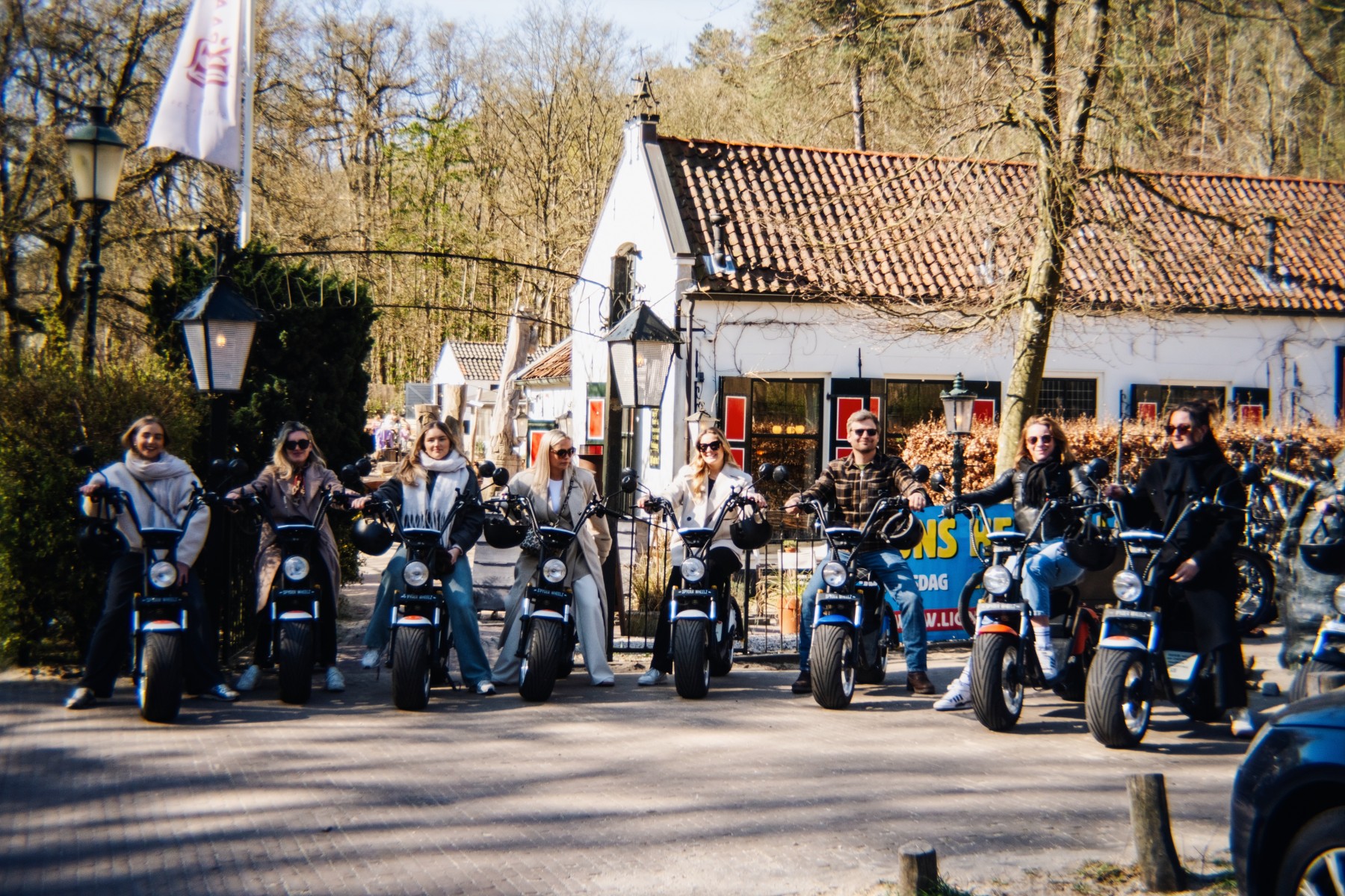 Groep mensen op elektrische scooters voor een wit huisje met rode dakpannen, omgeven door bomen.