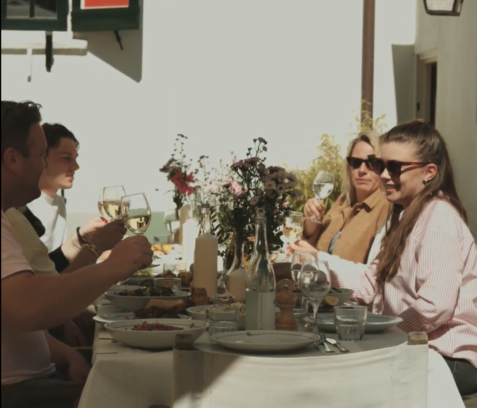 Groep vrienden geniet van een zonnige lunch buiten, omringd door bloemen en glazen witte wijn.
