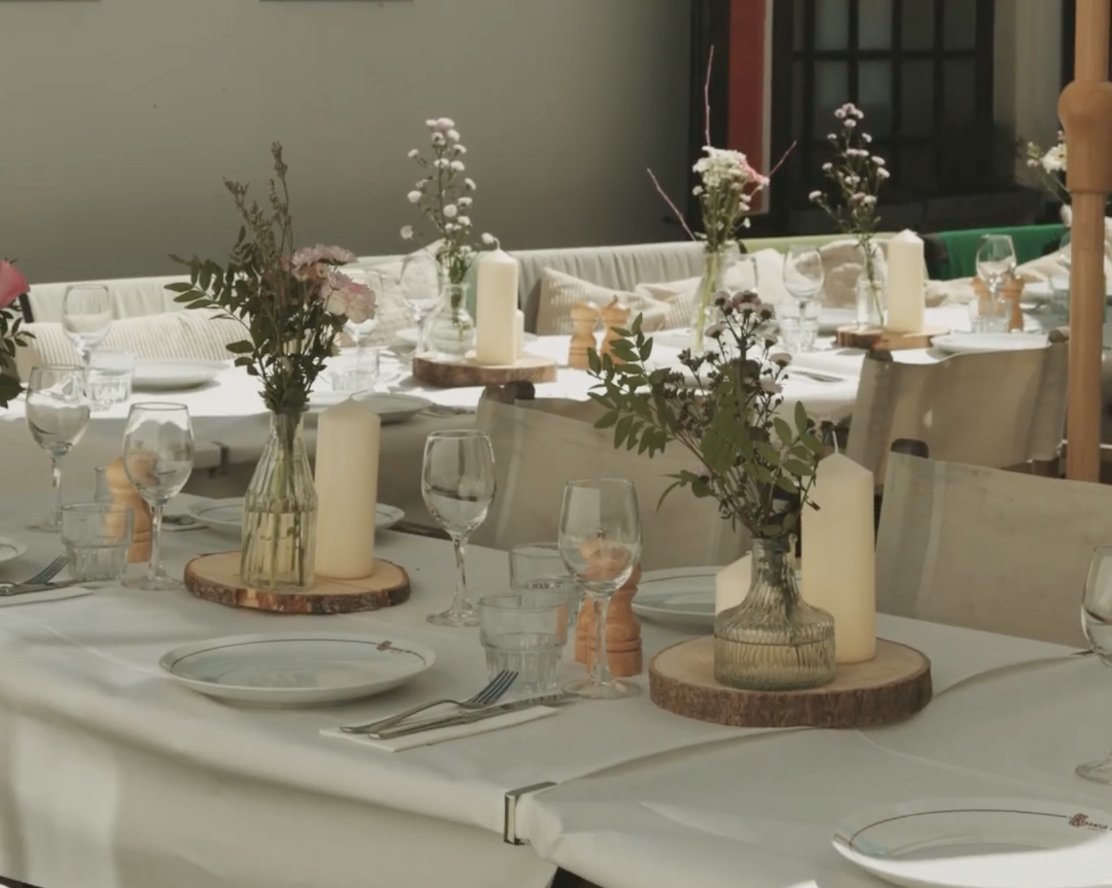 Elegante tafels met witte tafellakens, glazen vazen met bloemen, kaarsen en gedekte borden in een lichte ruimte.