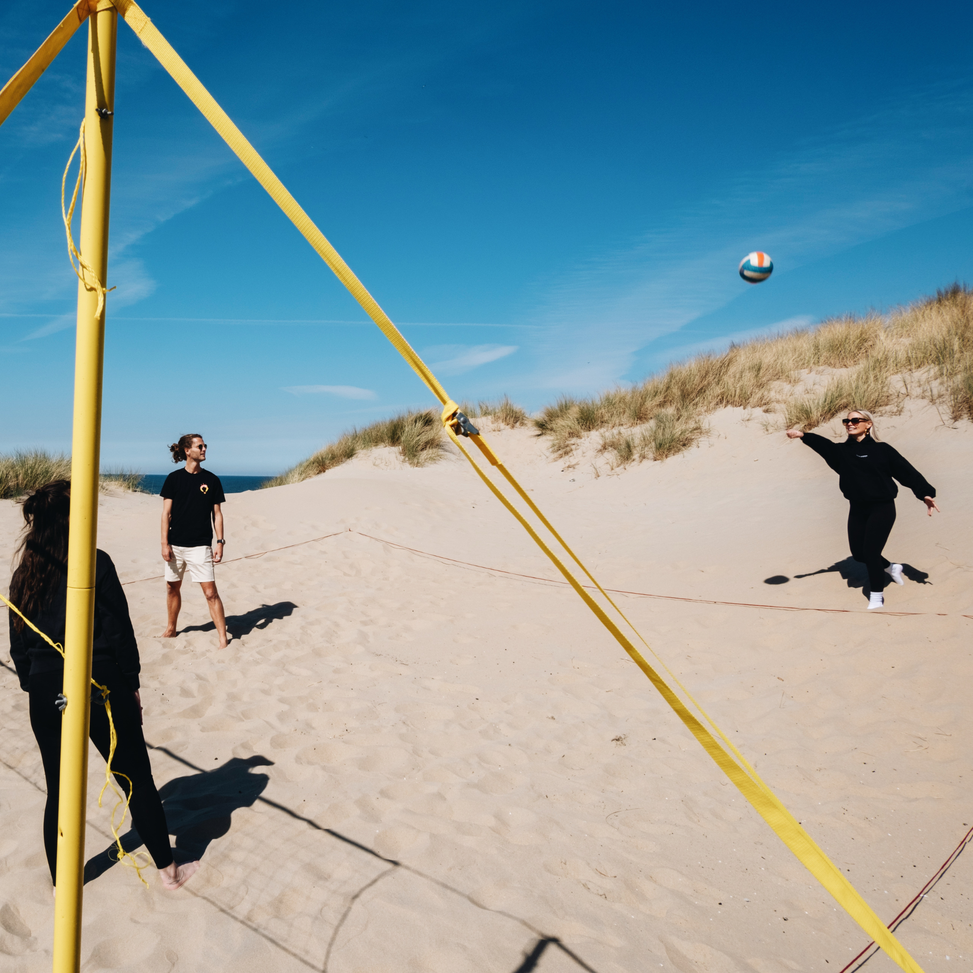 Mensen spelen beachvolleybal op een zonnig strand met duinen en blauwe lucht op de achtergrond.