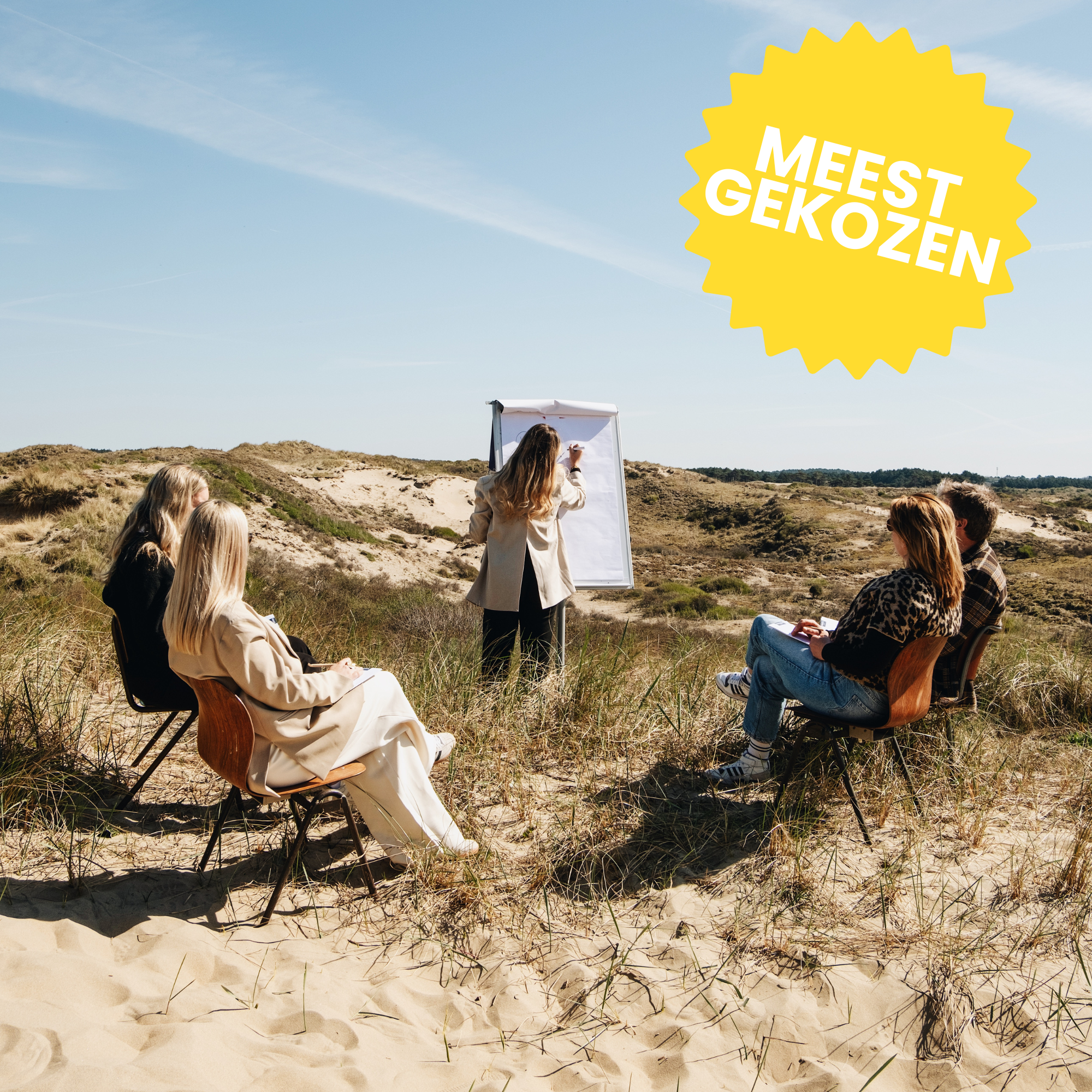 Groep mensen in een zandduinlandschap, zittend op stoelen, terwijl iemand op een flip-over schrijft.