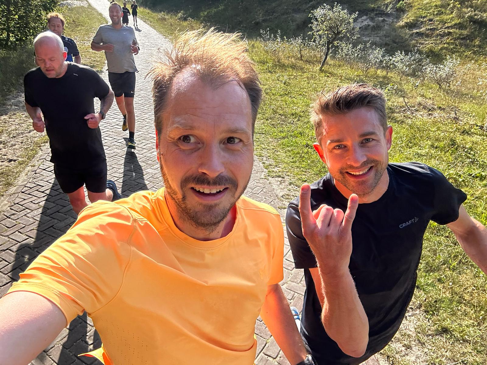 Mannen rennen op een zonnig pad in sportkleding, omgeven door groen gras en bomen.