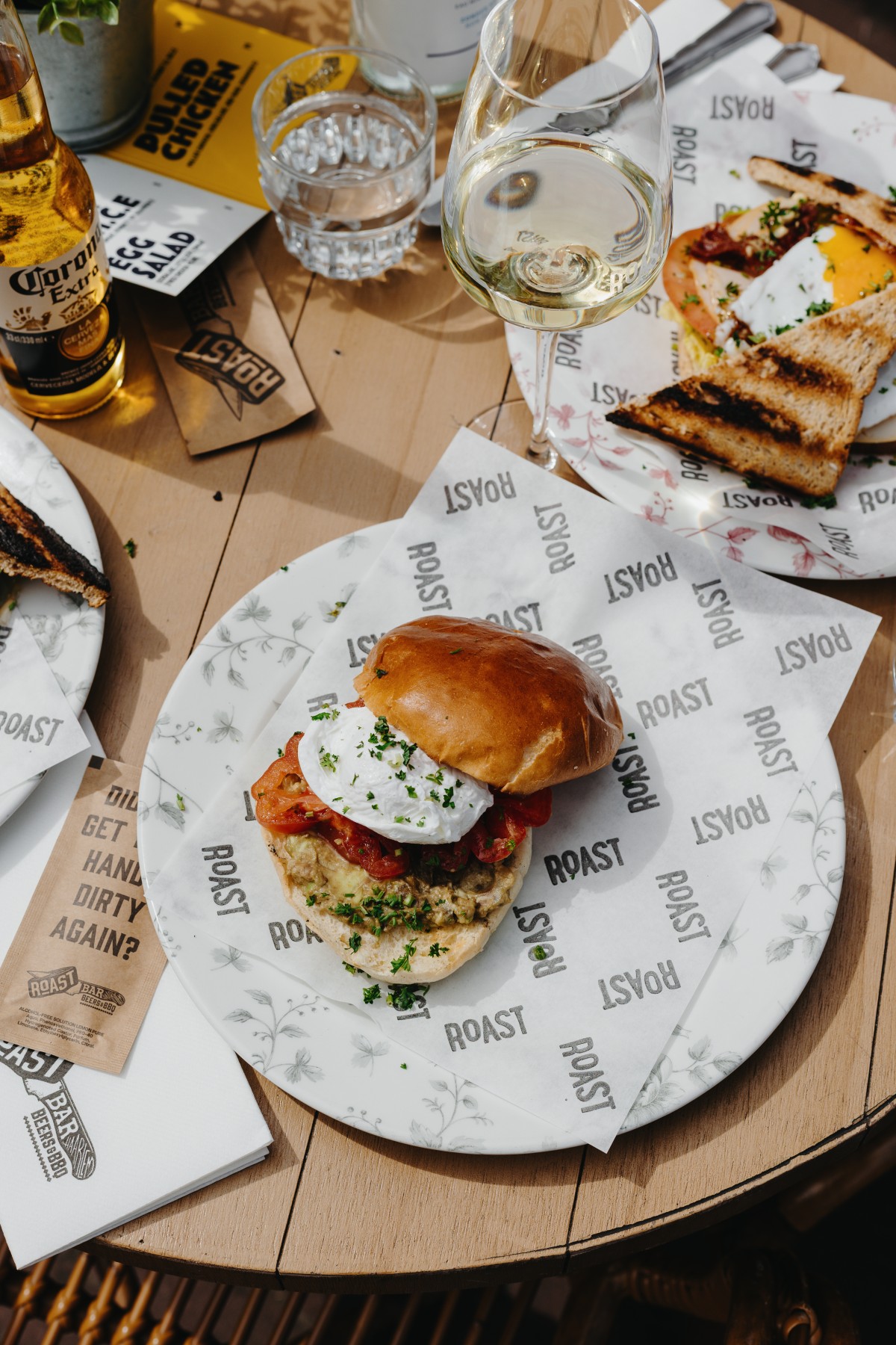 Hamburger met gepocheerd ei en tomaat op wit bord, omgeven door toast, glas witte wijn en flesje Corona.